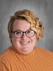 Smiling woman with curly hair wearing glasses and an orange sweater, against a gray background.