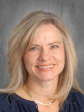 Smiling woman with light brown hair wearing a navy blouse and a necklace, set against a gray background.