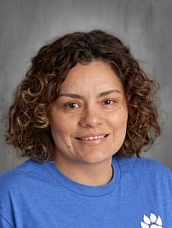 A person with curly hair smiles, wearing a blue shirt, against a blurred gray background.