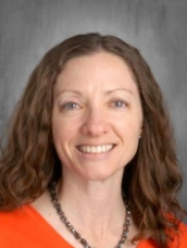 A woman with curly brown hair smiles at the camera, wearing an orange top and a necklace, against a gray background.