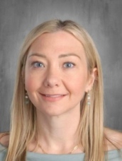 A woman with long blonde hair and blue eyes smiles against a gray background, wearing earrings and a light blue top.