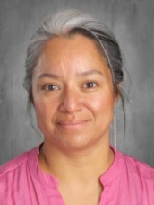 A woman with gray hair and a pink blouse smiles warmly at the camera against a gray background.