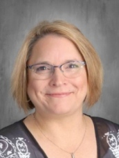 Portrait of a smiling woman with shoulder-length blonde hair and glasses, wearing a black patterned top.