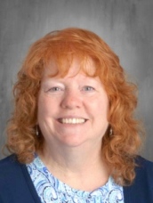Smiling woman with wavy orange hair wearing a blue top and a light blue patterned blouse against a gray background.