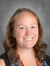 Headshot of a smiling woman with long, curly brown hair wearing a black top and colorful necklace against a gray background.