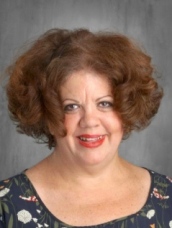 A woman with curly brown hair and a floral blouse smiles at the camera against a gray background.