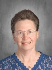 Woman with short brown hair, wearing glasses and a blue lace top, smiling against a gray background.