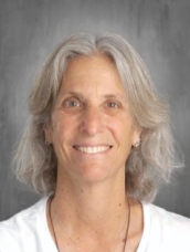 Smiling woman with shoulder-length gray hair, wearing a white shirt, against a gray background.