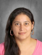A woman with long dark hair and hoop earrings wearing a pink shirt, smiling against a gray background.
