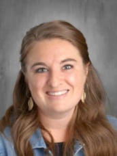A smiling woman with long brown hair and earrings, wearing a denim jacket, against a neutral background.
