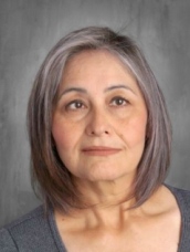 A woman with shoulder-length gray hair and a neutral expression, wearing a gray top, against a gray background.