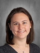 Smiling woman with shoulder-length brown hair, wearing a dark sweater against a gray background.