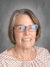 A middle-aged woman with short gray hair and glasses smiles, wearing a patterned blouse and pearl necklace.