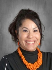 Smiling woman with dark hair in a black top wearing a vibrant orange beaded necklace, against a gray background.