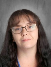 A woman with long, dark hair and glasses poses against a gray background. She has a calm expression.