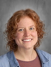 A smiling person with curly red hair, wearing a denim jacket over a striped shirt, against a gray background.