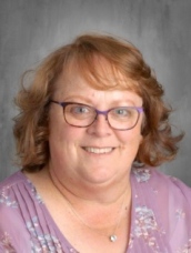 A smiling woman with curly auburn hair and glasses, wearing a floral purple blouse, poses for a portrait.