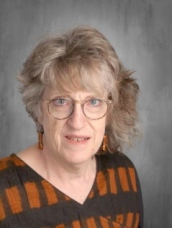A woman with curly gray hair and glasses, wearing a patterned brown and orange top, smiles in a neutral background.