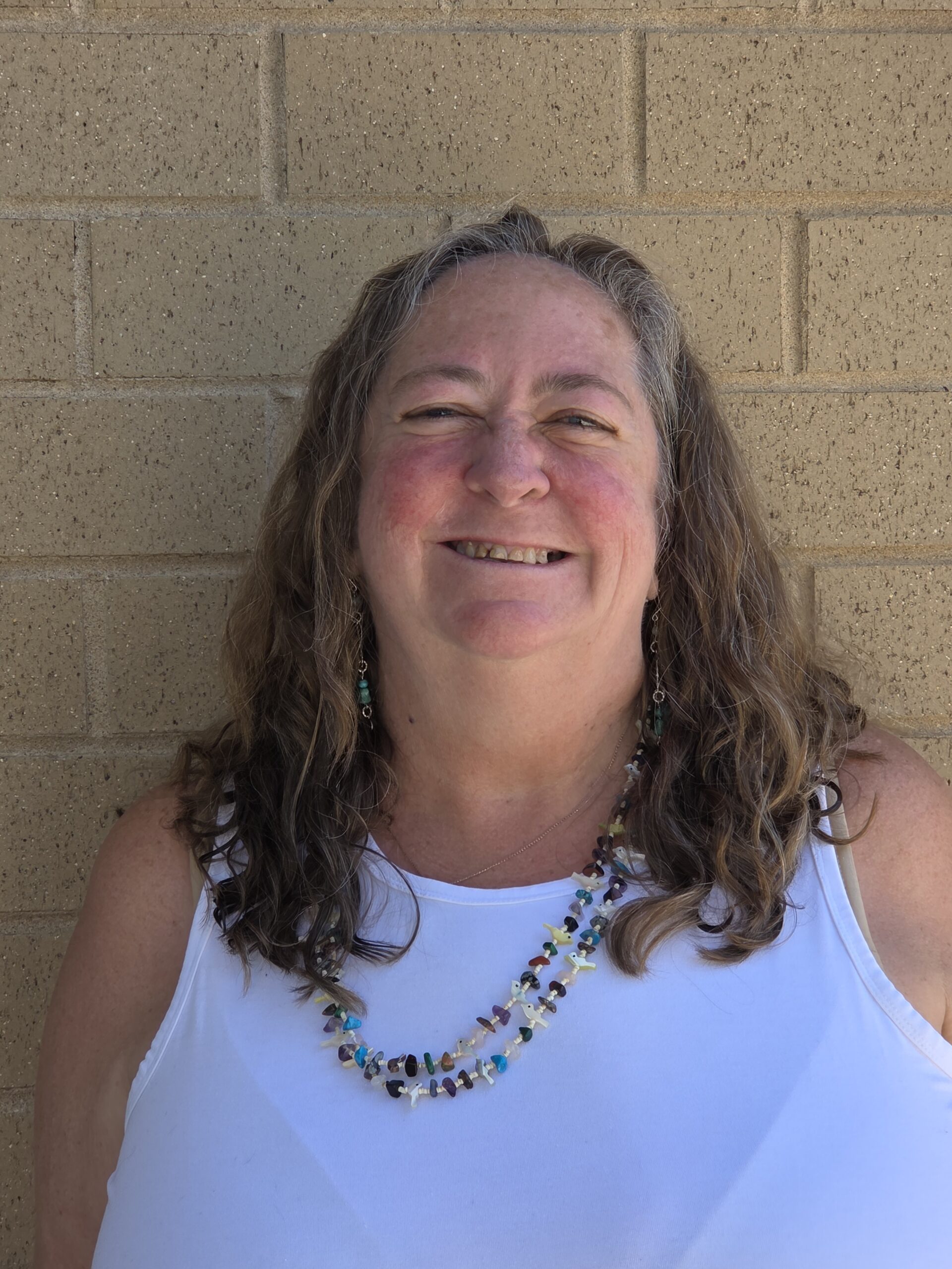 A smiling woman with curly hair wears a colorful beaded necklace against a beige brick background.
