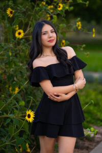 A young woman in a black off-shoulder dress poses among sunflowers, showcasing a natural outdoor setting.