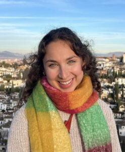 A woman with curly hair smiles warmly, wearing a colorful scarf, with a scenic city view in the background.