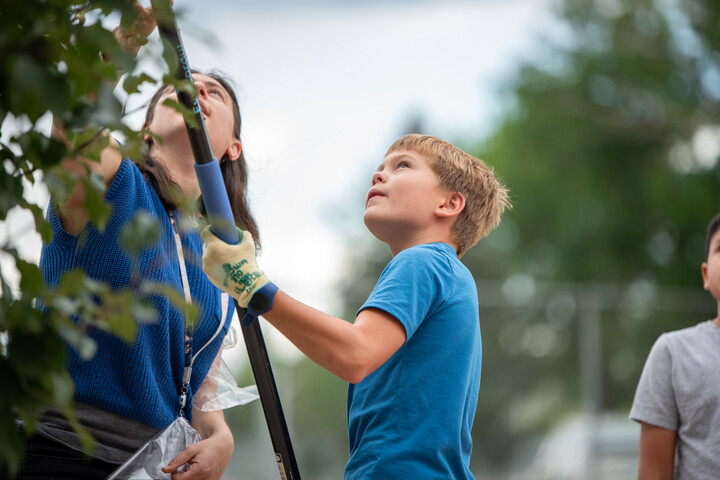 A child and an adult are working together to trim plants, with focused expressions and gardening tools in hand.