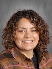 Smiling woman with curly hair wearing a plaid shirt, set against a neutral background.