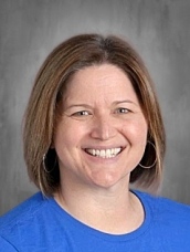 Smiling woman with shoulder-length brown hair, wearing a blue shirt and hoop earrings, against a gray background.