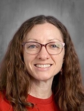 A smiling woman with long, curly brown hair and glasses, wearing a red shirt, poses for a professional photo.