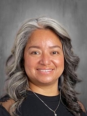 A woman with long, wavy gray hair smiles warmly at the camera, wearing a black top and a necklace.