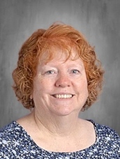Smiling woman with curly red hair wearing a blue floral top, set against a neutral background.