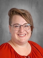A smiling person with short, styled hair and glasses, wearing a red blouse, against a grey background.