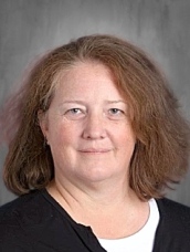 A woman with shoulder-length brown hair and a slight smile, wearing a black top, against a gray background.