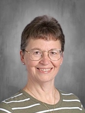 Smiling woman with short hair and glasses, wearing a striped shirt, against a gray background.