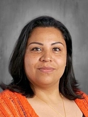 A woman with dark hair and a warm expression, wearing an orange top, poses for a portrait against a plain background.