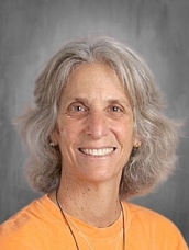 Smiling woman with wavy gray hair, wearing an orange shirt, against a gray background.
