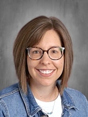 A smiling woman with short brown hair and glasses, wearing a denim jacket over a white shirt, against a gray background.