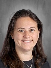 Smiling woman with long brown hair, wearing a black shirt, set against a gray background.