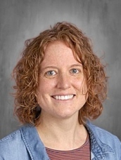 Smiling woman with curly red hair, wearing a denim jacket over a striped shirt, against a gray background.