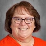 Smiling woman with brown hair and glasses, wearing an orange sweater, against a neutral gray background.