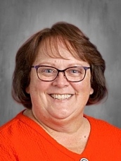 Smiling woman with brown hair and glasses, wearing an orange sweater, against a neutral gray background.