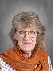 A woman with curly hair and glasses, wearing an orange scarf, smiles against a gray background.