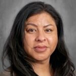 A woman with long black hair, wearing a neutral-colored top, smiles against a gray background.