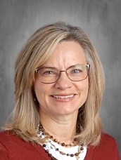 A smiling woman with long, light brown hair, glasses, and a necklace, set against a gray background.