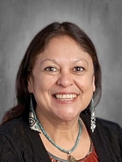 Smiling woman with long dark hair, wearing earrings and a necklace, set against a neutral background.