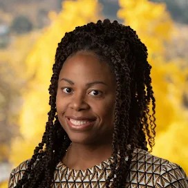 A woman with braided hair smiles, wearing a patterned top, with a backdrop of yellow autumn leaves.